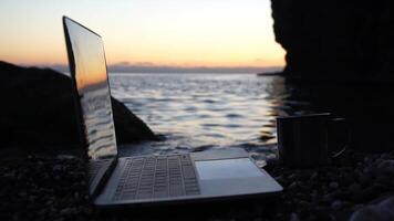 Silhouette of laptop and cup on the beach over sea sunset. Sparkling sea water on background. Freelancer, blogger working on notebook on the beach. video