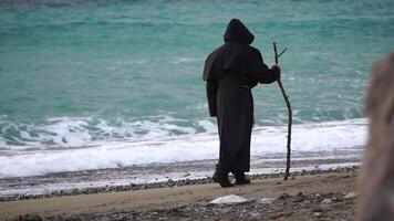 Monks wearing black robes walks on the beach with wooden stick in hands at morning. Unrecognizable person, slow motion. video