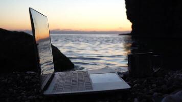 Silhouette of laptop and cup on the beach over sea sunset. Sparkling sea water on background. Freelancer, blogger working on notebook on the beach. video