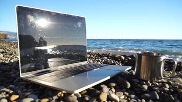 Silhouette of laptop and cup on the beach over sea sunset. Sparkling sea water on background. Freelancer, blogger working on notebook on the beach. video