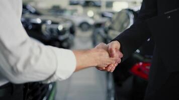 Close-up shot of a confident man in a white shirt shaking hands with an assistant in a business suit in a car dealership. Happy and mutually beneficial between two people in a car showroom video
