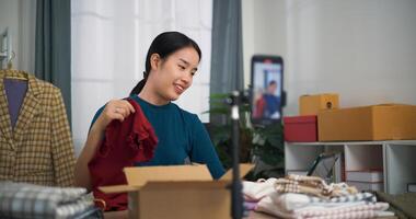 Portrait of young Asian business owner selling clothes and chatting with customers via live broadcast on mobile phone, online shopping photo