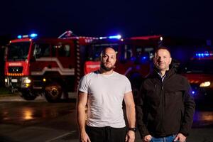 Group of firefighters, dressed in civilian clothing, stand in front of fire trucks during the night, showcasing a moment of camaraderie and unity among the team as they reflect on their duties and the photo