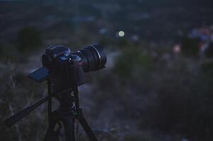 A black digital semi-mirror camera photographic equipment, with movable mockup touch screen, placed on tripod in nature, capturing steady picture or recording of the city in the night and dawn. photo