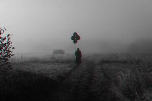terrible man in a hooded cloak with balloons in his hand stands in a foggy field photo