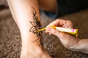 A henna artist carefully applies a intricate floral design to a womans ankle using a cone-shaped applicator. The design is already partially complete, and the henna artist is adding more detail to the delicate pattern. photo