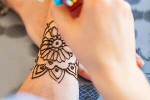 This image shows a woman applying a henna tattoo to another womans hand. The womans hand is resting on a table with a pineapple patterned tablecloth. The henna tattoo is a intricate design of flowers and swirls. photo