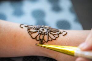 This image shows a henna artist applying an intricate floral design on someones forearm. The artist is using a traditional henna cone, and the design is in progress. The image captures the detail and artistry involved in the henna process. photo