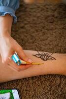 A woman is applying a henna tattoo on her lower leg using a cone. She is wearing a blue shirt and has her leg resting on a brown rug. The tattoo is a simple flower design. photo