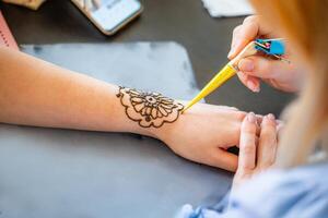 A woman is applying henna to a clients wrist. The clients arm is resting on a table. The woman is using a cone to apply the henna, which will create a temporary tattoo design. photo