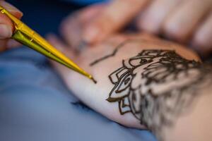 A close-up image of a woman receiving a henna tattoo on her wrist. The artist is using a cone filled with henna paste to meticulously draw an intricate design on the womans skin. The background is a fabric with a pattern of pineapples. photo