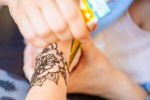 A close-up image of a woman receiving a henna tattoo on her wrist. The artist is using a cone filled with henna paste to meticulously draw an intricate design on the womans skin. The background is a fabric with a pattern of pineapples. photo