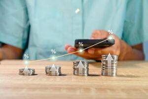 Man using a smartphone to calculate the financial interest rate hikes with coins stacked on wooden table photo