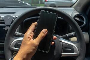 Male hand holding a smartphone with a car steering wheel in the background. Driving while using a smart phone concept. photo