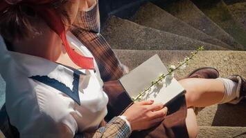 A girl sits sideways on the steps of the university and writes in a notepad. She holds a blooming white branch in her hands. Retro image in the sun's rays photo