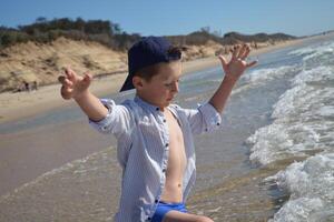 A todler boy in a cap and shirt on the ocean coast enjoys the sea, waves and vacations. The joy of a child at the sight of waves on the ocean. photo