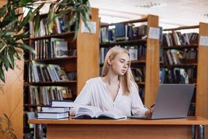 Attractive Multiethnic Student Making a STEM Class Homework in a Textbook, While Researching Quiz Answers on a Laptop Computer. Young Blonde Female Studying in a Public Library with Classical Interior photo
