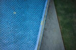 an aerial view of a swimming pool with a checkered tile pattern. The pool is surrounded by a concrete deck and a grassy area or plants or flower area. The scene is calm and inviting. photo