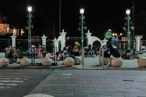 a group of people sitting on benches in kilometer nol, yogyakarta and round stone structures in a public square at night. The scene is peaceful and relaxed, with a sense of community and togetherness photo