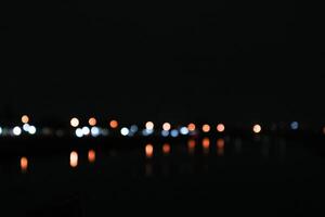 blurred view of a cityscape at night, with the lights of buildings reflecting in a body of water. The scene is dark and mysterious, with the lights providing a sense of warmth and life photo