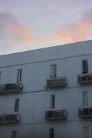 white apartment building with several windows and balconies. The sky is a soft pink and purple at sunset, suggesting a beautiful sunset. The scene is peaceful and serene photo