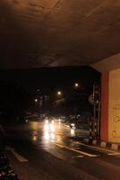 a wet street at night, with a traffic light and a few cars passing under a bridge. The street is illuminated by streetlights, and the reflection of the lights can be seen in the wet pavement photo
