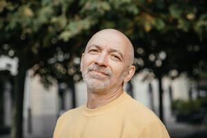 Senior man with a gentle smile, wearing a yellow sweater, standing under the shade of trees in an urban environment photo