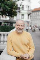 Cheerful bald senior man in a yellow sweater, leaning on a stone balustrade in an urban environment, radiating happiness photo