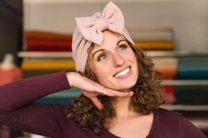 Joyful fashion designer playfully posing with a handmade pink bow headband, her hand as a pretend support, in her vibrant fabric-lined studio photo