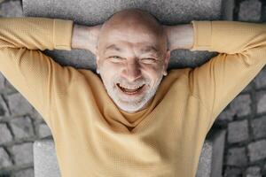 Playful bald senior man lying on his back on an urban bench, making funny faces, capturing a moment of levity photo