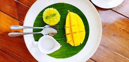 Flat lay of Thai local dessert food with mango, sticky rice, jug of coconut milk, spoon and fork on banana leaf in white plate on wooden table with copy space at cafe. Delicious and Natural material. photo