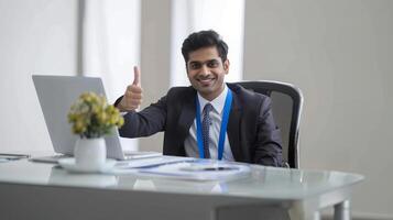 A happy Indian professional man gives a thumbs up while sitting at his desk with a laptop in an office photo