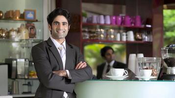 A confident Indian businessman stands at a coffee shop smiling and looking at the camera photo