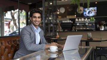 A confident Indian businessman enjoys a coffee at a trendy cafe while working on his laptop photo