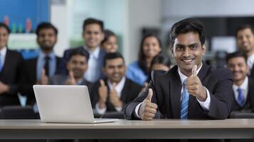 A happy Indian businessman gives a thumbs up while sitting at a table with a laptop photo