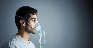 A young Indian man using a nebulizer for relief from breath issues on a grey background photo