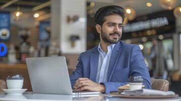 A confident Indian businessman enjoys a coffee at a trendy cafe while working on his laptop photo