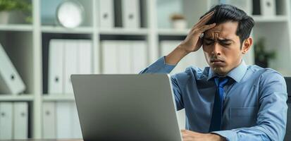 An Indian business man sitting in front of laptop computer holding his forehead while feeling pain from stress at the office photo