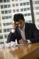 A young Indian sad man in formal attire is sitting at an office desk with crumpled papers photo
