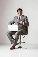 A smiling Indian man in sits on a bar chair with a laptop giving a thumbs up photo