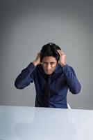 A middleaged Indian man sitting at a white table holding his head in pain as if he is feeling so sad photo