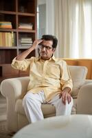 An Indian man sitting on a sofa in a living room and looking sad while touching his forehead in thought photo