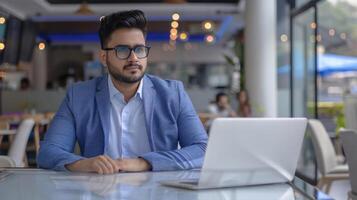A confident Indian businessman enjoys a coffee at a trendy cafe while working on his laptop photo