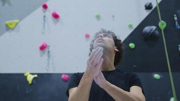 Young climber preparing with chalk, looking up towards the climbing wall with colorful holds in an indoor gym, showcasing focus and determination before a climb video