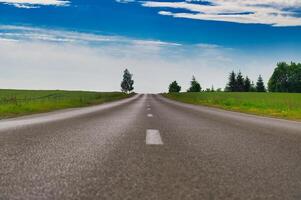 Empty asphalt road leading into horizon under blue sky with clouds and green fields photo