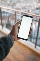 A hand holding a smartphone with a blank screen on a balcony with a city view in the background photo