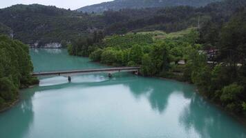 Drone view of turquoise mountain lake and bridge across it. emerald lake lies nestled in rocky terrain and dense forest. bridge spans lake, showcasing tranquil lake, bridge and surrounding nature video