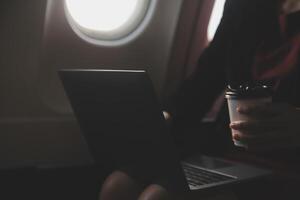 Blonde female tourist checking incoming notification on smartphone sitting on seat of airplane with netbook.Young businesswoman share media from telephone on laptop computer during plane flight photo