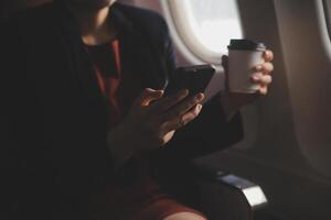 Blonde female tourist checking incoming notification on smartphone sitting on seat of airplane with netbook.Young businesswoman share media from telephone on laptop computer during plane flight photo