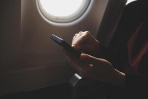 Blonde female tourist checking incoming notification on smartphone sitting on seat of airplane with netbook.Young businesswoman share media from telephone on laptop computer during plane flight photo